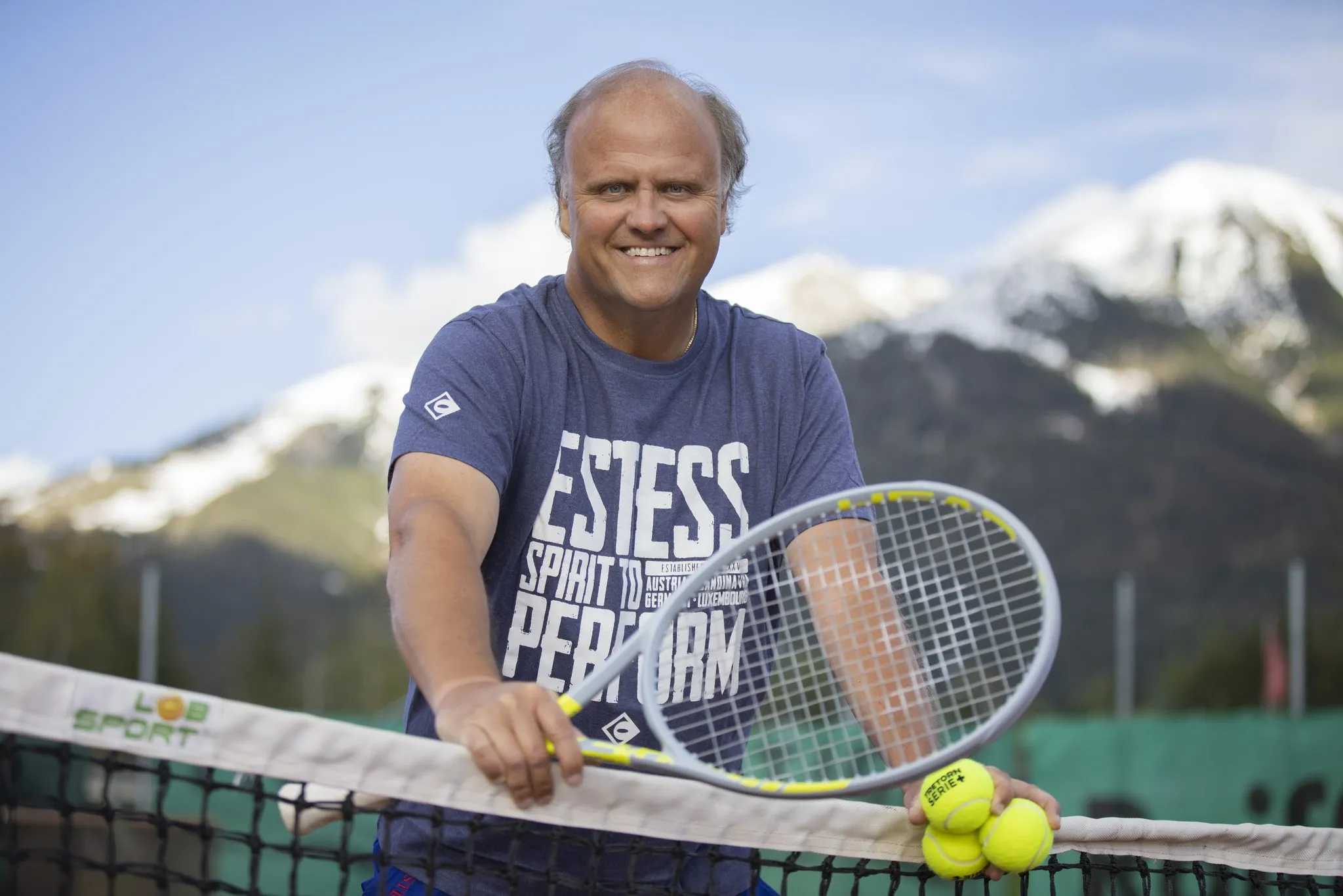 Tennistrainer mit Schläger und Bällen am Netz vor Bergkulisse, Portrait on Location für ESTESS