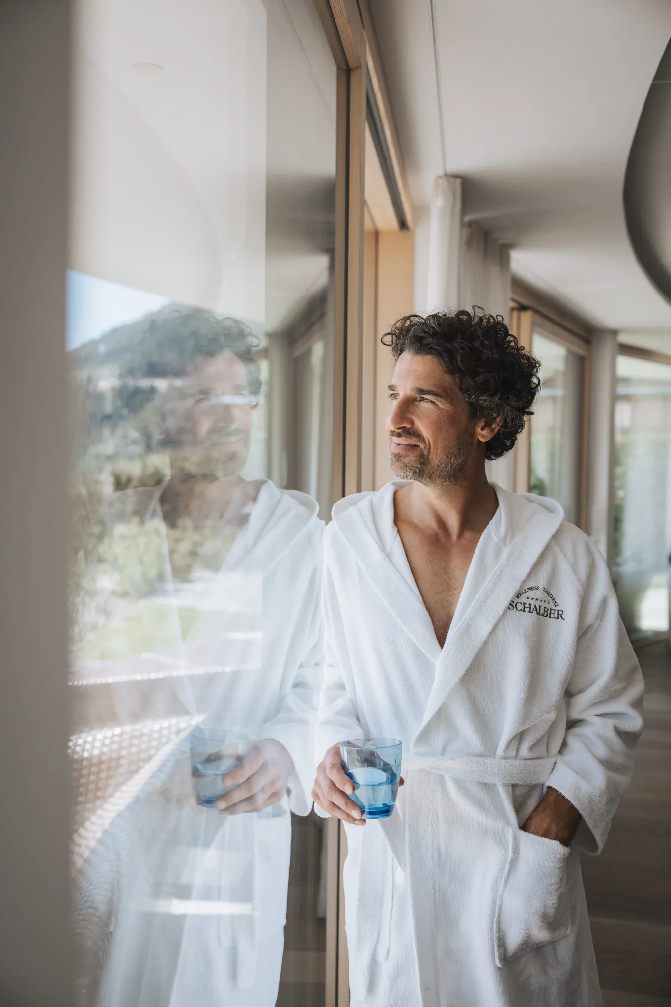 Mann im Bademantel mit Wasserglas am Panoramafenster, Wellnessresidenz Schalber in Serfaus